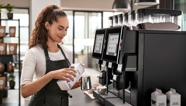 Weibliche Barista gießt vor einem Kaffeevollautomaten Milch in ein Kännchen.